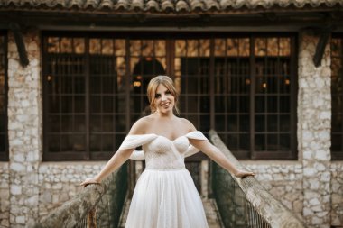 Güzel gelin portresi ahşap köprüde arkasında taş evle duruyor.