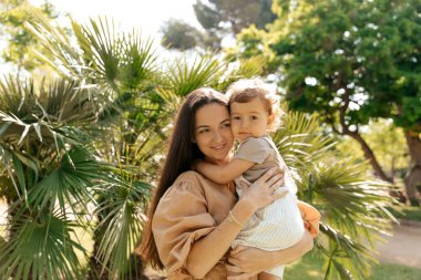 Güneşli ve sıcak bir günde, egzotik bitkilere karşı uzun siyah saçlı bir kadının kucağında çocuğunun olduğu açık hava fotoğrafını çek. Yüksek kalite fotoğraf