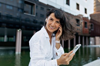 Kısa saçlı romantik bir kadın kablosuz kulaklıkla müzik dinliyor ve akıllı telefonuyla kameraya gülümsüyor. 