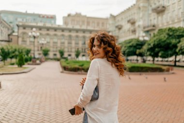 Kıvırcık saçlı, beyaz tişörtlü, çantalı ve akıllı telefonlu heyecanlı kızıl saçlı kadın güneşli bir akşamda şehirde yürüyor. Dişleriyle gülümseyen, rahat, beyaz bir kadının yakın çekimi..