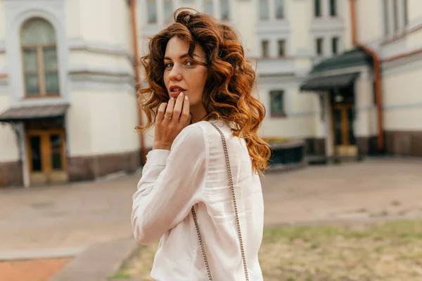 Romantic tender woman with curly hair wearing white blouse looking around at camera while walking in the city in warm sunny evening. Portrait of cheerful pretty woman looking away and smiling.