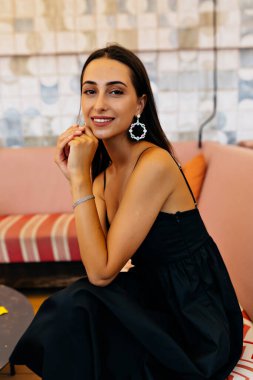 Siyah elbiseli, küpeli, koyu renk saçlı sevimli bir kadın kameraya poz veriyor. Gülümsüyor ve bir yaz terasında dinleniyor. 