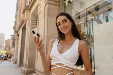 Beyaz bluzlu, bol siyah saçlı güzel bir bayan akıllı telefonuyla şarkı söylüyor ve şehir caddesinde kameraya gülümsüyor. Avrupalı güzel bir kızın yakın çekilen fotoğrafı açık havada vakit geçiriyor. 