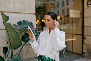 Siyah saçlı, beyaz tişörtlü ve parlak etekli çekici bir kadın şehirde yürüyor. Akıllı telefon kullanıyor ve kablosuz kulaklıkla müzik dinliyor.