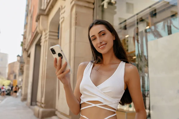 Beautiful lovely lady with loose long dark hair wearing white top is carolling smartphone and smiling at camera on city street .Close up photo of european good-looking girl spend time outdoor