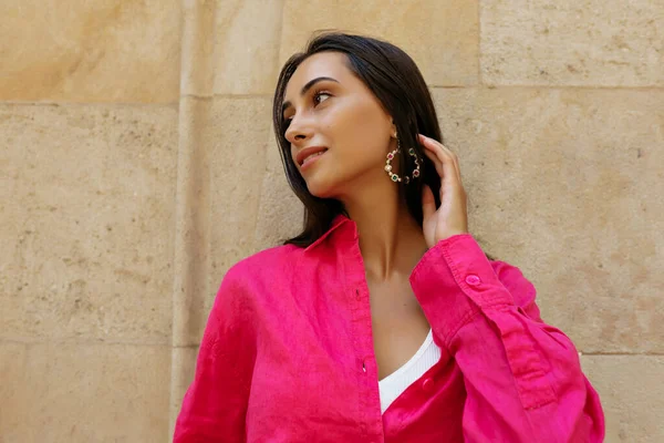 Close up profile photo of brunette girl with long loose hair wearing pink shirt is looking code and touching her hair while walking in the city and looking aside .