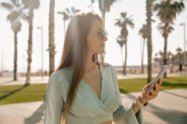 Uzun saçlı güzel bir kadının profil portresi. Akıllı telefon kullanıyor ve gün ışığında seti doldurup müzik dinliyor. Mavi gömlek ve güneş gözlüğü takıyor.