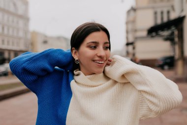 Çekici gülümseyen, koyu saçlı, harika gülümseyen, yana bakan ve ellerini kaldıran kadının yakın portresi. Mavi beyaz süveter giyiyor.