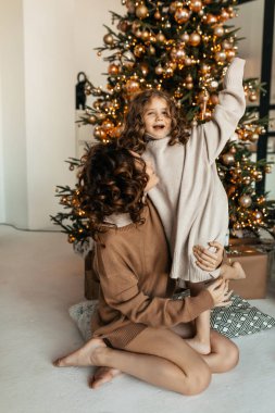Kıvırcık saçlı, süveter giyen, Noel ağacının önünde annesiyle oynayan komik, heyecanlı küçük kız. Tatil, yeni yıl, Noel, aile. 
