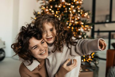 Kıvırcık saçlı güzel bir bayanın Noel ağacının önünde küçük kızıyla oynayıp vakit geçirdiği portreyi yakından çek.