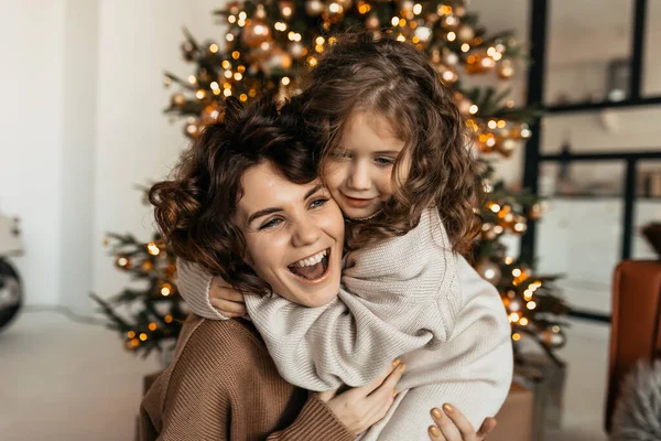 stock image Young woman in warm clothes playing with daughter on Christmas tree background. Indoor shot of two positive girls in sweaters hugging and smiling