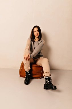 Koyu renk saçlı, bej örgü takım elbiseli ve izole edilmiş arka planda sandalyede oturan çizmeli modern bir kadının tam boy fotoğrafı. 