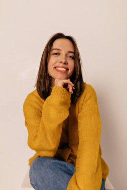 Kot pantolonlu, şık, sarı gömlekli, genç, mutlu, esmer kadın. İçtenlikle gülümsüyor, kafasını kaldırıp beyaz odada yere oturuyor..