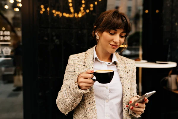 charming pretty european woman with dark hair wearing light jacket and blouse drinking coffee and using smartphone. Happy excited dark-haired woman laughing and walking around city.
