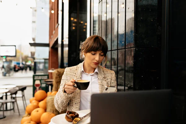 Stylish pretty woman with dark hair wearing light blouse and jacket sitting on open air cafe with coffee and working on laptop.