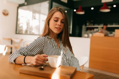 Kapalı alanda, uzun saçlı, çizgili tişörtlü, güneşli bir kafede oturan kahve ve kruvasanın keyfini çıkaran çekici bir kızın portresini çek..