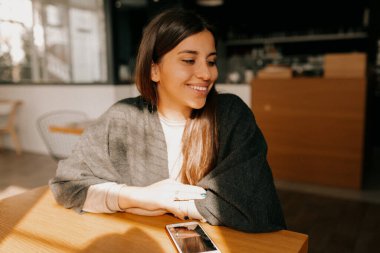 Akıllı telefon kullanan gülümseyen bir kadın ve kafenin penceresinde. Kapalı alanda gülümseyen mutlu kızın portresi kafeteryada sabah kahvesinin tadını çıkarıyor.. 