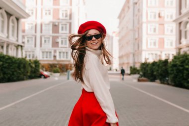 Saçları açık, güneş gözlüğü takan, kırmızı bereli ve etekli sevimli bir bayanın yakın portresi kameraya doğru dönüyor, gülümsüyor ve sokakta zıplıyor. 