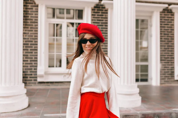 Happy excited smiling girl with loose hair wearing red beret and red skirt smiling, having fun and walking down the street on background of modern buildings