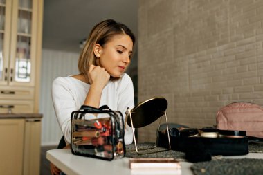 Güzel makyaj sanatçısı küçük bir aynaya bakarken makyaj yapıyor. Güzel sarışın kadın aynaya bakıp makyaj yapıyor ve makyaj malzemelerini fırçayla sürüyor..