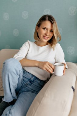 Sarı kısa saçlı, beyaz gömlekli, kot pantolonlu, kanepeye oturmuş kahveyle keyfi yerinde güzel bir kadın. Sıcak ve güneşli bir günde evde dinlenen güzel bir kızın kapalı bir fotoğrafı..