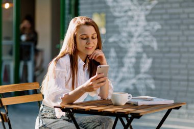 Açık havada oturan beyaz gömlek ve kot giyen gevşek saçlı romantik, sevimli kız. Akıllı telefonu olan ve gülümseyen bir kafe. Tarz sahibi kız açık kafede arkadaşlarını bekliyor. 