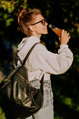 Beyaz gömlekli, kot pantolonlu, güneşli bir sabahta ders çalışmaya giden ve meyve suyu içen hoş bir bayan öğrenci.. 