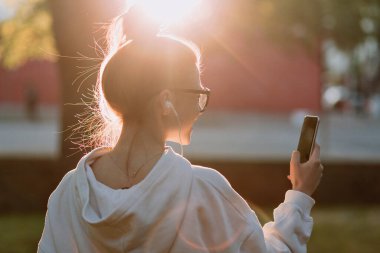 Güneş gözlüklü, beyaz tişörtlü, güneş gözlüklü ve güneşli bir sabahta kulaklıkla müzik dinleyen çekici bir genç kızın portresini yakından çek. 