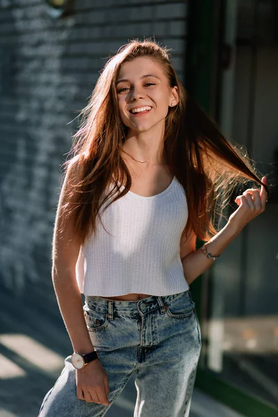 Cute smiling happy girl with loose hair and wonderful smile posing at camera while standing outside against modern city cafe in sunlight