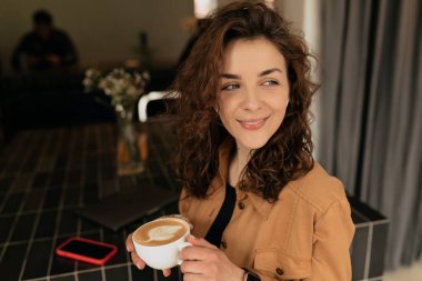 Çekici, gülümseyen, harika gülüşlü, pofuduk saçlı ve elinde bir fincan kapuçino tutan güzel bir kadının portresi. 