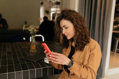 Şık saçlı, akıllı telefon kullanan ve şehir kafesinde dinlenen sevimli bir bayan. Kahverengi gömlekli hoş bir kız kafede kahve içerken telefonda sohbet ediyor.