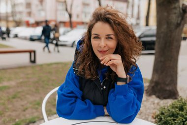 Güzel, tüylü saçlı, mavi gömlekli, koyu renk ceketli bir kadın. Açık havada, şehir kafesinde otururken kamerayı zehirliyor..