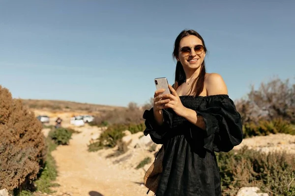 Charming cute woman with happy wonderful smile wearing black elegant dress and sunglasses using phone and traveling on road with mountains and ocean on background