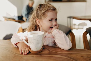 Close up portrait of smiling happy girl with wavy hair sitting in cafe in sunny cafe and enjoying weekend day with mother. Close up photo of happy smiling kid with cacao 