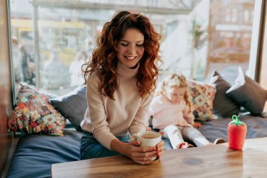 Attractive smiling good-looking red-haired woman resting in sunny modern cafe with her little adorable daughter. Happy family spend weekend in cafe and enjoying cacao. 