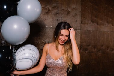Romantic cute lady with long light hair wearing festive dress looking down and touching hair while standing with balloons over dark backgrounds 