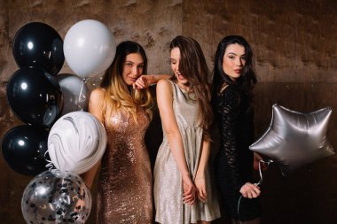 Adorable three female models in festive elegant dresses posing with balloons on dark background. Girlfriends with flowing hair wear shiny dresses. Friendship concept.