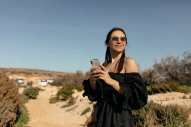 Attractive adorable woman with dark hair wearing black dress and sunglasses using smartphone and looking aside with happy smile on background of island nature 