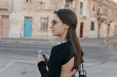 Close up portrait from back of charming pretty brunette girl were black top walking in old city. She were sunglasses and using smartphone 