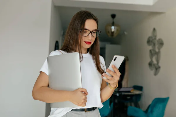 Adorable cute modern woman with dark hair and red lipstick wearing white t-shirt I using smartphone and holding laptop ion background of modern apartment