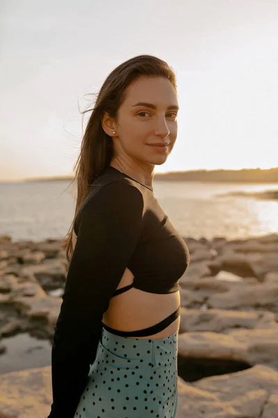 Outdoor portrait of brunette stylish girl with loose dark hair wearing black top and skirt posing at camera with happy smile on background of beautiful beach on sunset.