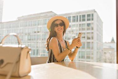 Pretty charming woman with wonderful smile wearing hat and sunglasses sitting on summer terrace with city view and scrolling smartphone 