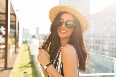 Come up portrait of charming smiling woman with dark loose hair wearing sunglasses and hat turn around to camera in sunlight on blur city background