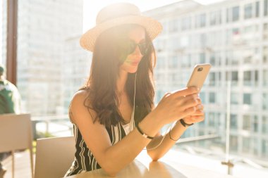 Close up portrait of stylish charming brunette girl with wavy hair wearing hat and sunglasses using smartphone and listening music in headphones 