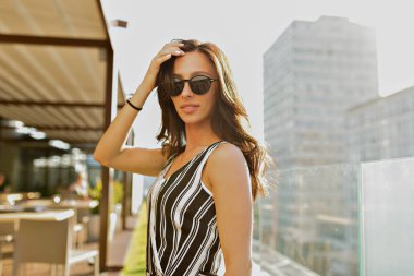 Close up outdoor photo of brunette stylish girl with wonderful smile wearing sunglasses and striped summer t-shirt is posing to camera and touching her hair while walking in the city 