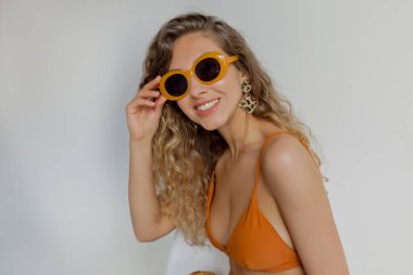 well-looking compelling girl with snow-white smile and long wavy hair wearing orange glasses and orange swimsuit posing to camera over isolated background.