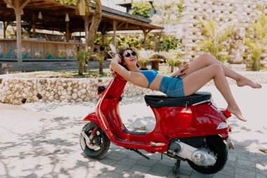 Smiling adorable stylish woman in swimsuit and shorts reading outdoor on the red bike in summer sunny evening. Happy girl having fun and resting on background of exotic plants