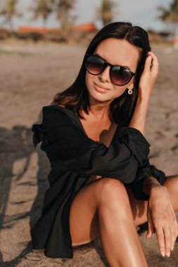 Stylish cute charming woman with dark hair wearing black shirt posing to camera with adorable smile and looks to camera on sandy beach
