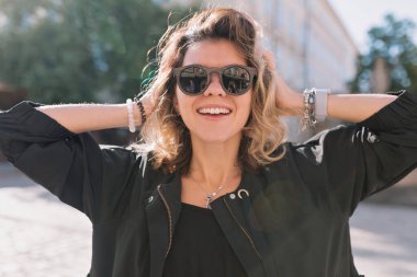 Close up portrait of adorable happy girl with wonderful smile posing to camera while wearing sunglasses and black jacket on background of city square in sunlight 