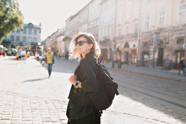 Happy adorable pretty woman with curtly hairstyle wearing black jacket and sunglasses with bag pack walking outdoor in sunlight with happy smile 